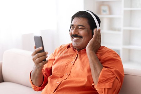 Photo for Happy mature indian man with moustaches using smartphone and wireless headphones, have video call with family, watching movie online, sitting on couch at home, copy space. Digital world - Royalty Free Image