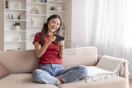 Foto de Joven coreana alegre jugando videojuegos en el teléfono inteligente mientras se relaja en el sofá, mujer asiática feliz usando auriculares inalámbricos descansando en el sofá en la sala de estar, disfrutando de las tecnologías modernas - Imagen libre de derechos