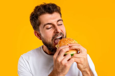 Téléchargez les photos : Mangeur de malbouffe. Gros plan de l'homme affamé savourant un hamburger savoureux, prendre une bouchée de burger à emporter sur fond de studio jaune. Équilibre nutritionnel entre alimentation et fringales - en image libre de droit