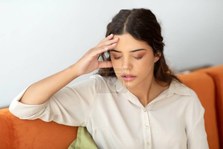 Photo for Headache Concept. Stressed young lady suffering from migraine at home, sick millennial woman touching temple while sitting on couch in living room, feeling unwell, having acute pain, closeup shot - Royalty Free Image