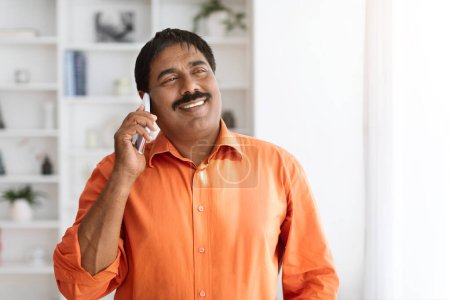 Téléchargez les photos : Gai mature indien homme avec moustache porter smart casual avoir conversation téléphonique et sourire, en regardant l'espace de copie vierge pour la publicité, posant au bureau à la maison - en image libre de droit
