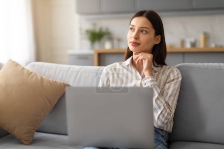 Photo for Pensive young woman in striped shirt using laptop computer while sitting comfortably on grey couch, hand on chin and looking aside at free space - Royalty Free Image