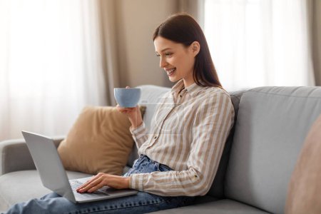Photo for Smiling relaxed young woman enjoying cup of coffee while casually working on her laptop computer or surfing internet, sitting on gray sofa, free space - Royalty Free Image
