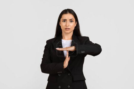 Focused young european businesswoman in black suit making time out sign with her hands, displaying serious expression, standing against grey background