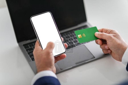 Téléchargez les photos : Homme d'affaires tenant un smartphone avec un écran blanc dans une main et une carte de crédit verte dans l'autre, prêt à effectuer un paiement en ligne, avec un ordinateur portable en arrière-plan - en image libre de droit