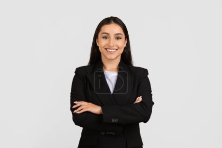 Photo for Professional young european businesswoman with beaming smile, dressed in stylish black suit, confidently stands with arms crossed against grey background - Royalty Free Image