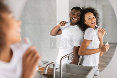 Foto de Alegre pareja afroamericana cantando posando cerca del espejo con cepillos de dientes, disfrutando de la rutina de cuidado dental juntos y divirtiéndose, en el baño moderno en casa. Enfoque selectivo - Imagen libre de derechos