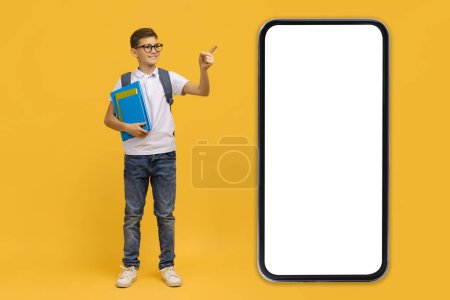 Photo for Educational App. Excited Teen Schoolboy Pointing At Huge Smartphone With Blank Black Screen, Male Kid Wearing Eyeglasses Holding Workbooks, Recommending New Application On Yellow Background, Mockup - Royalty Free Image