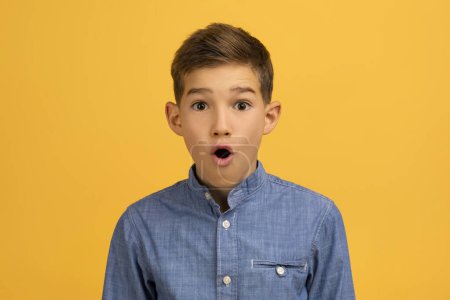 Photo for Astonished teenage boy with wide-eyed expression posing against yellow studio background, shocked teen kid wearing casual blue denim shirt capturing moment of surprise, looking at camera - Royalty Free Image