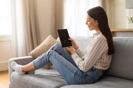 Photo for Happy young female in casual striped shirt lounging on gray sofa while browsing on digital tablet, looking at blank screen, place for mockup and design - Royalty Free Image