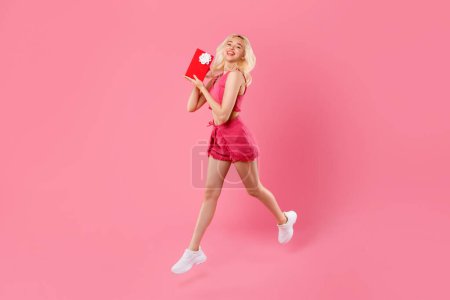 Photo for Blonde young woman in pink, caught mid-leap with beaming smile, clutching red gift box with white bow, embodying pure joy against pink backdrop - Royalty Free Image