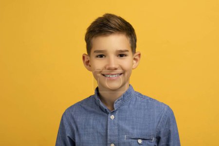 Photo for Happy teen boy with charming smile wearing casual blue denim shirt posing against vibrant yellow studio background, cheerful male teenage kid exuding youthful energy and confidence, copy space - Royalty Free Image
