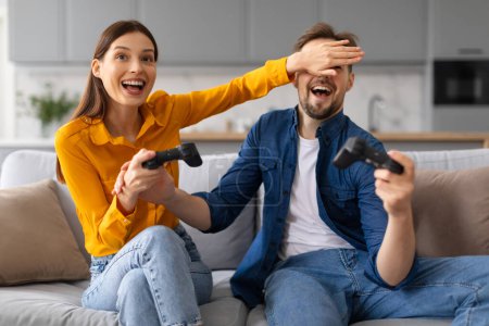 Téléchargez les photos : Joyeux jeunes conjoints engagés dans des jeux vidéo, avec la femme riant couvrant les yeux des hommes, créant une atmosphère ludique à la maison, assis sur le canapé - en image libre de droit