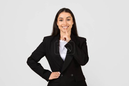 Photo for Young businesswoman in sleek black suit makes shush sign with her finger to lips, suggesting confidentiality or secret, posing on grey background - Royalty Free Image