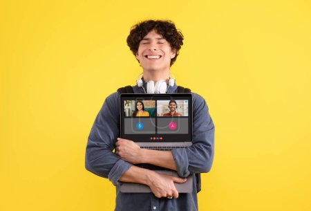Photo for A young man with curly hair and headphones around his neck is smiling contentedly while embracing a laptop showing an active video call on a yellow background. Meeting, social networks - Royalty Free Image