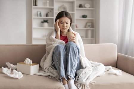 Photo for Unwell young asian woman wrapped in blanket suffering from headache at home, sick korean female feeling unwell, sitting on couch with pained expression, surrounded by tissues, copy space - Royalty Free Image