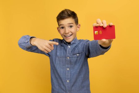 Téléchargez les photos : Sourire adolescent garçon pointant avec confiance à la carte de crédit rouge dans sa main, adolescent garçon heureux représentant la responsabilité financière ou l'éducation des consommateurs, debout sur fond jaune vif - en image libre de droit