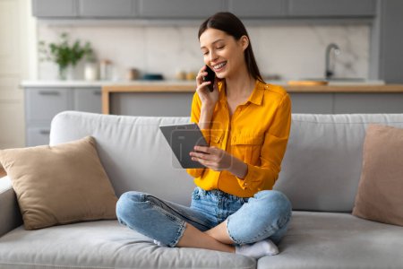 Photo for Cheerful young woman sitting cross-legged on sofa, engaged in conversation over her phone while simultaneously browsing on digital tablet - Royalty Free Image