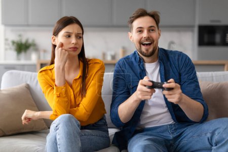 Foto de Hombre absorto sosteniendo el controlador de juego y jugando videojuegos con emoción mientras la mujer escéptica se sienta a su lado, reflexionando sobre su compromiso - Imagen libre de derechos