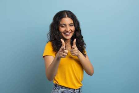 Téléchargez les photos : Jeune étudiante indienne positive pointant des deux mains vers la caméra et souriant, posant isolée sur fond bleu studio coloré, espace de copie. Concept des gestes - en image libre de droit