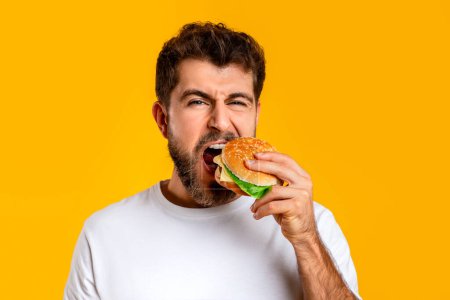 Photo for Hungry Guy Taking Bite Of Tasty Burger On Yellow Studio Backdrop, Closeup Portrait. Funny Young Bearded Man Eating Delicious Cheeseburger With Appetite. Concept Of Nutrition And Cheat Meals - Royalty Free Image