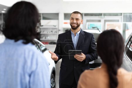 Photo for A professional car salesman in a well-tailored suit confidently discusses vehicle options with a young couple at a spacious and modern car dealership, digital tablet in hand. - Royalty Free Image