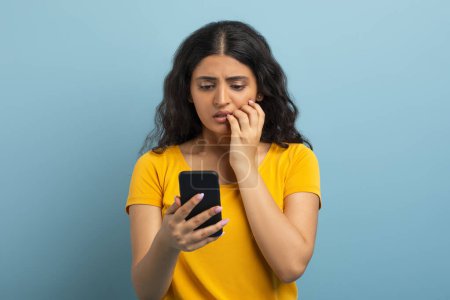 Photo for Confused pretty curly young indian woman wearing yellow t-shirt using cell phone isolated on blue studio background, reading weird news or text message, touching her face - Royalty Free Image