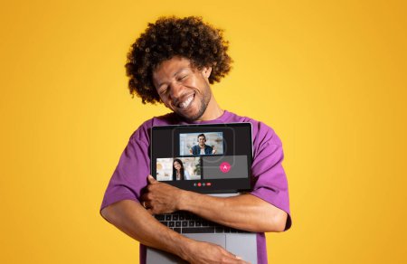 Téléchargez les photos : Un homme aux cheveux bouclés étreint joyeusement son ordinateur portable en montrant une vidéoconférence avec des interactions positives entre les participants sur un fond jaune vif. Réunion, réseaux sociaux - en image libre de droit