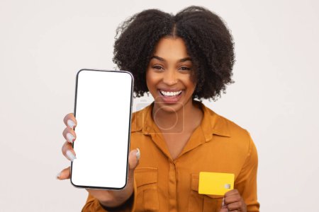 Foto de Encantada mujer afroamericana milenaria con el pelo rizado que muestra un teléfono inteligente con una pantalla en blanco y una tarjeta de crédito, posiblemente promoviendo la banca en línea o servicios de compras - Imagen libre de derechos