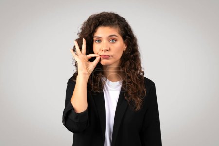 Photo for Young european businesswoman in black suit making silence gesture with her finger on her lips, signaling the need for quiet or secrecy, looking at camera on grey backdrop - Royalty Free Image