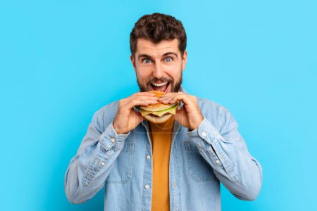 Photo for Happy caucasian man experiencing delight of juicy cheeseburger, biting delicious takeaway meal on blue backdrop, studio portrait shot. Concept of tasty versus healthy, cheat meals and cravings - Royalty Free Image