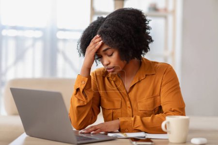 Foto de La triste y estresada mujer afroamericana milenaria con una blusa de mostaza muestra signos de frustración o dolor de cabeza mientras trabaja en una computadora portátil, con una taza de café en el escritorio en la oficina - Imagen libre de derechos