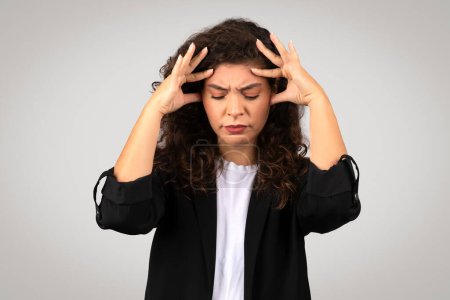 Foto de Mujer de negocios joven y estresada con expresión de dolor en la cara, sosteniendo su cabeza en frustración o dolor de cabeza, vistiendo chaqueta negra y camisa blanca, posando sobre fondo gris - Imagen libre de derechos