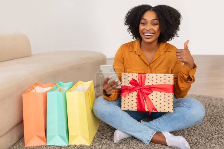 Foto de Feliz millennial mujer afroamericana con una amplia sonrisa sosteniendo un regalo y su teléfono inteligente, dando un pulgar hacia arriba mientras está sentado en el suelo cerca de coloridas bolsas de compras en casa - Imagen libre de derechos