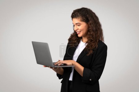 Foto de Joven empresaria comprometida centrada en escribir en el portátil, demostrando un flujo de trabajo eficiente y conectividad de pie sobre un fondo gris - Imagen libre de derechos