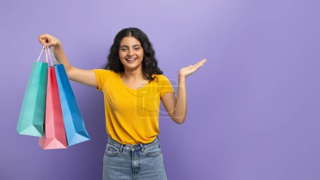 Great offer, season sale. Happy young indian woman student carrying colorful paper bags purchases and showing blank copy space for advertisement on purple studio background. Shopping concept