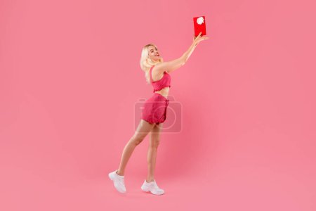 Photo for Exuberant blonde in playful coral outfit joyously tossing red gift box in the air, full of vibrant energy, against bright pink background, full length - Royalty Free Image