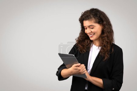 Photo for Young european businesswoman in black suit using digital tablet, reviewing important documents or chatting online with clients, posing on gray studio background, free space - Royalty Free Image