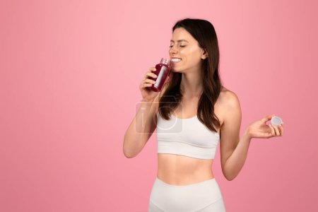 Foto de Una mujer europea consciente de la salud en ropa deportiva blanca disfruta de un jugo de remolacha fresca mientras sostiene una tapa de botella, colocada sobre un fondo rosa, que encarna un estilo de vida saludable - Imagen libre de derechos