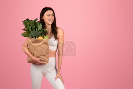 Téléchargez les photos : Une joyeuse femme européenne en tenue de fitness rit tout en tenant un sac en papier rempli de fruits et légumes frais, symbolisant une alimentation saine et un mode de vie sur fond rose - en image libre de droit