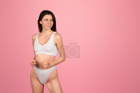 Photo for A smiling european young woman with sleek brunette hair looks to the side with a thoughtful expression, wearing a simple white swimsuit against a plain pink backdrop, studio - Royalty Free Image