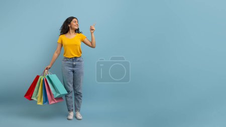 Tolles Angebot, Saisonverkauf. Fröhliche junge Studentin aus dem Osten trägt bunte Papiertüten bei sich und zeigt auf leere Kopierflächen für Werbung auf blauem Studiohintergrund. Einkaufskonzept