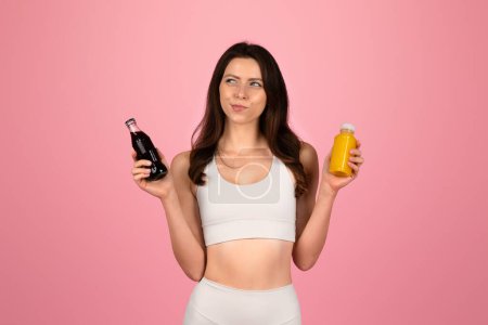 Photo for A contemplative young woman in a white tank top and leggings holds a bottle of soda in one hand and a juice in the other, pondering a choice against a soft pink background - Royalty Free Image
