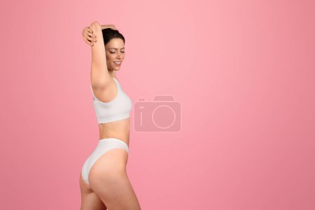 Photo for Content and composed, european young woman with closed eyes raises her arms above, showcasing a serene moment in a snug white sports top and bikini on a pink background, studio - Royalty Free Image