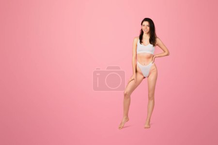Photo for A radiant happy young woman with long brown hair poses confidently in a white two-piece swimsuit against a vibrant pink background, exuding fitness and health, full length - Royalty Free Image