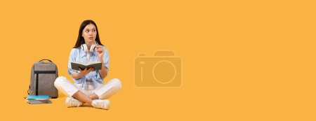 Téléchargez les photos : Jeune femme pensive en chemise et casque tenant le livre, avec sac à dos et cahiers à côté d'elle sur fond orange vif, panorama avec espace libre - en image libre de droit