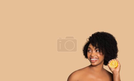 Téléchargez les photos : Femme afro-américaine rayonnante tenant une tranche de citron frais à côté de son visage, souriant joyeusement, représentant les bienfaits naturels des soins de la peau sur fond beige, espace libre, bannière - en image libre de droit