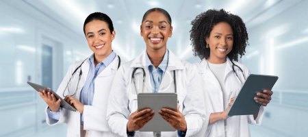 Téléchargez les photos : Trois femmes médecins souriantes en blouse blanche et chemises bleues, tenant des tablettes numériques, se tiennent ensemble dans un couloir d'hôpital, respirant compétence et convivialité - en image libre de droit