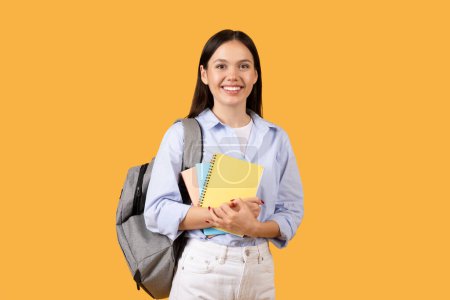 Photo for Happy european female student in blue shirt holding notebooks, wearing grey backpack, standing at camera against plain yellow background - Royalty Free Image