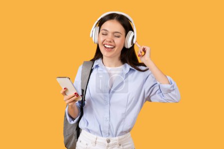 Foto de Joven encantadora con auriculares disfrutando de la música desde su teléfono, sonriendo y bailando, con mochila gris sobre fondo amarillo - Imagen libre de derechos
